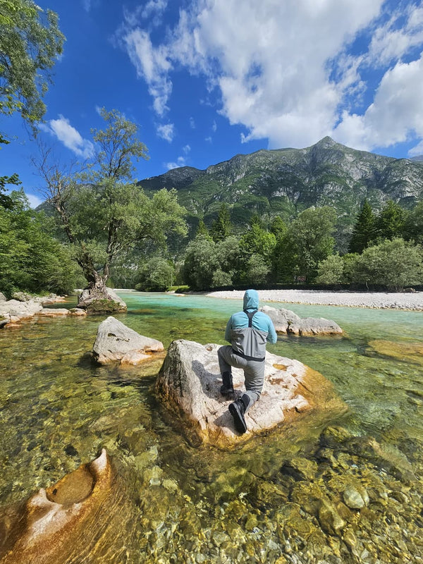 Person in teal hooded jacket and gray waders kneeling on pale rock in clear turquoise Krka River with trees and mountains