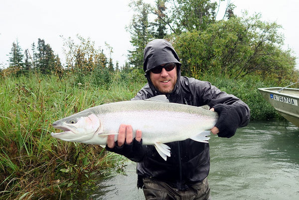 Angler in black jacket and gloves holding large silver steelhead over river beside a boat on a fishing trip