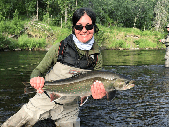 Angler in beige waders and green jacket holding silvery trout in shallow river with forest — Mission Lodge fishing trip