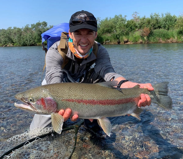 Angler holding a large rainbow trout in clear creek, wearing gray fishing jacket and black cap on Mission Lodge fishing trip