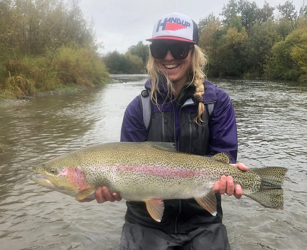 Smiling angler in purple waterproof jacket and trucker hat holding large iridescent rainbow trout on a river fishing trip