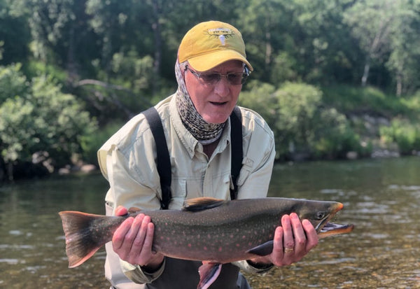 Angler in yellow cap and tan shirt holding dark-spotted trout over shallow river, green trees, Kulik Lodge fishing trip
