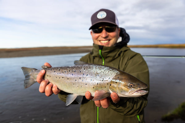 Angler in olive waterproof jacket and dark cap holding a shiny silver trout with black spots on a river fishing trip