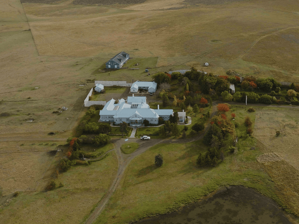 Aerial view of pale blue lodge with light metal roofs, garden, pond and autumn trees surrounded by open grassland