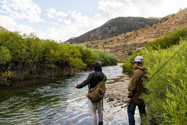 Two anglers in olive and black waterproof jackets and waders fly fishing on a mountain river shore, casting rods