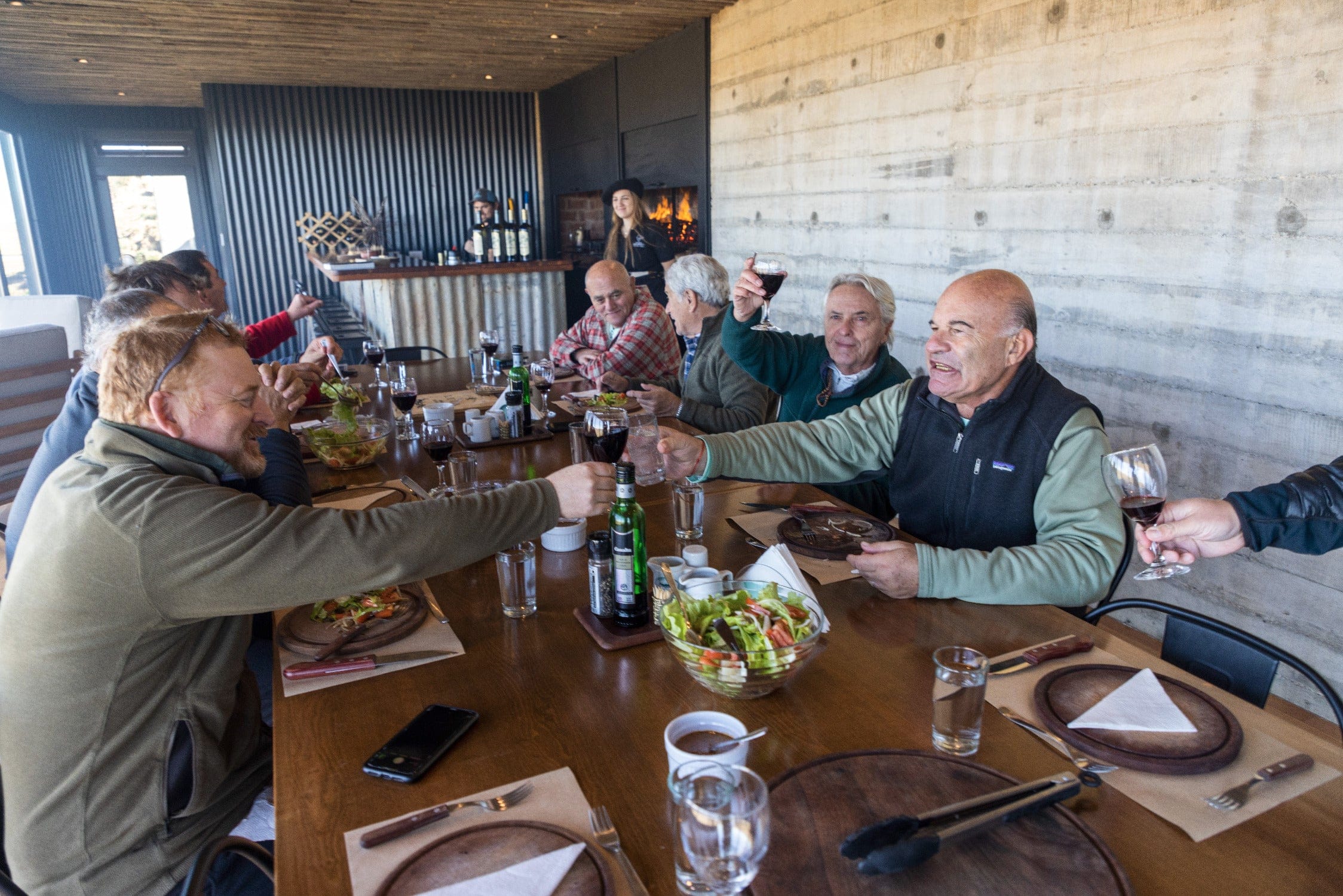 People toasting red wine at a long wooden dining table in rustic Maria Behety lodge with concrete wall and corrugated bar