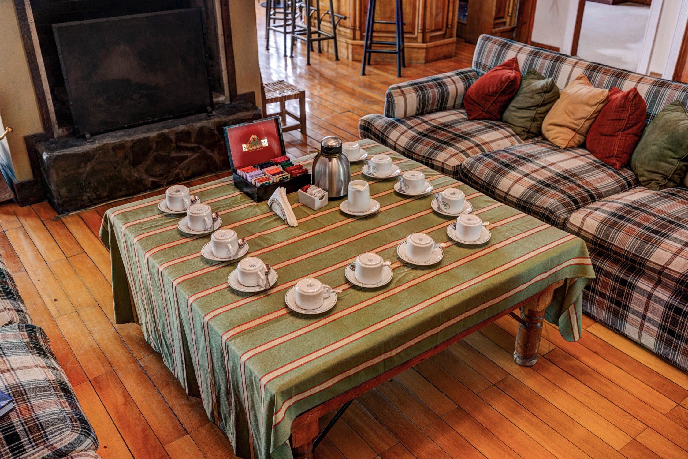 Cozy lodge sitting area with green striped tablecloth, white teacups on saucers, stainless thermos and plaid sofa cushions