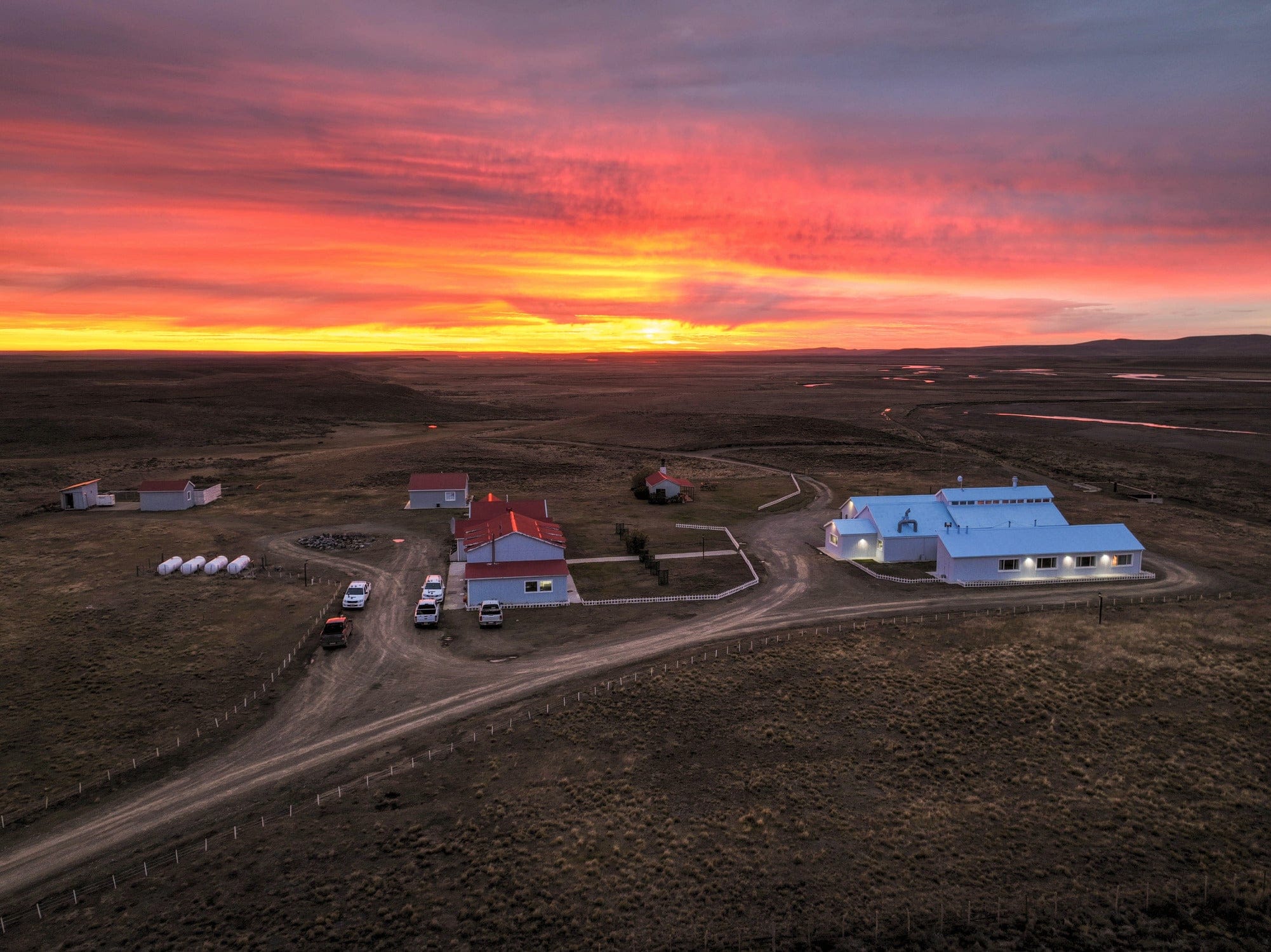 Maria Behety Lodge red-roof cabins and white metal barn on windswept plain under vivid pink-orange sunset - remote scenery