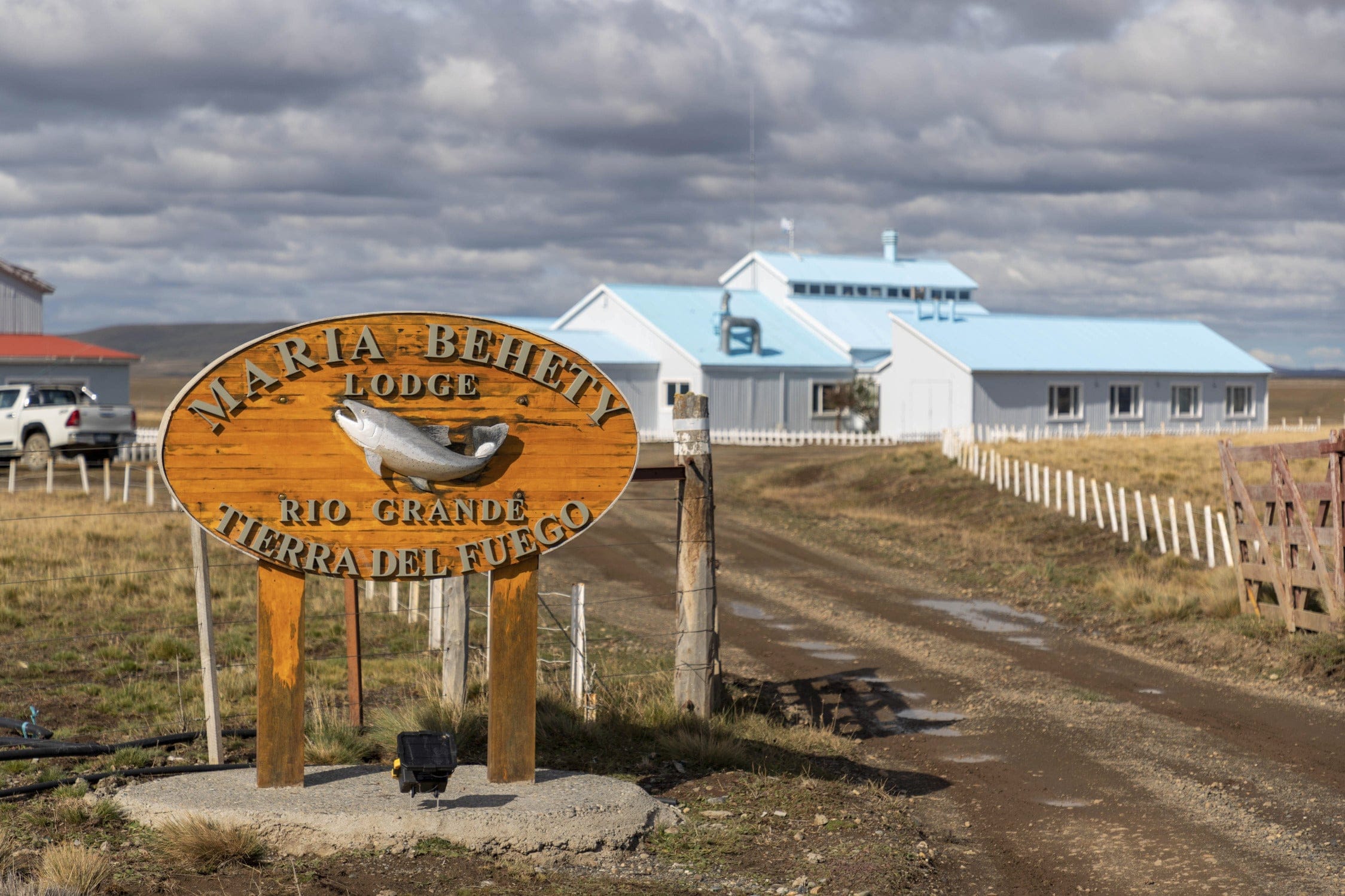 Wooden Maria Behety Lodge sign with carved fish, blue metal-roof lodge and dirt road under cloudy sky