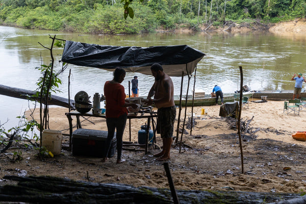 Black tarp camp kitchen on sandy Rewa riverbank with people cooking, moored boats and dense green forest, river camp trip