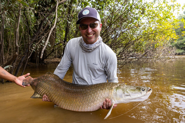 Smiling angler in gray sunshirt and trucker cap holding huge bronze-scaled arapaima in muddy river, Rewa River Camps fishing