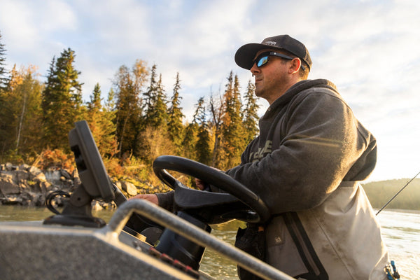Man in gray hoodie and black cap steering an aluminum boat on a river with evergreen shore - fishing trip scene
