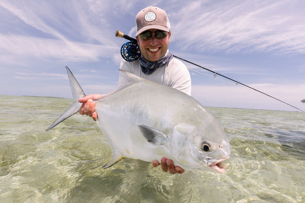 Smiling angler in tan Trident cap holding a large silver permit fish in clear green water on a fly fishing trip