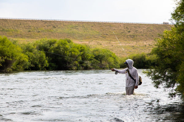 Fly fisherman in silver gear casting rod in river with lush green banks and rolling hills backdrop