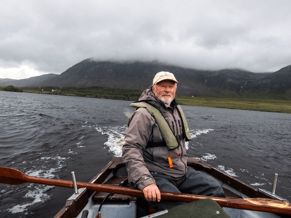 Bearded guide in beige cap and grey jacket in wooden boat on dark lake with misty mountains, fly fishing Ireland