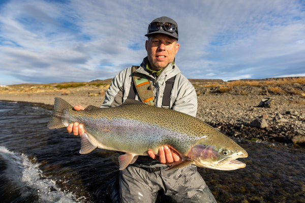 Angler in gray jacket and cap holding large speckled rainbow trout over rocky lake shore during fishing trip