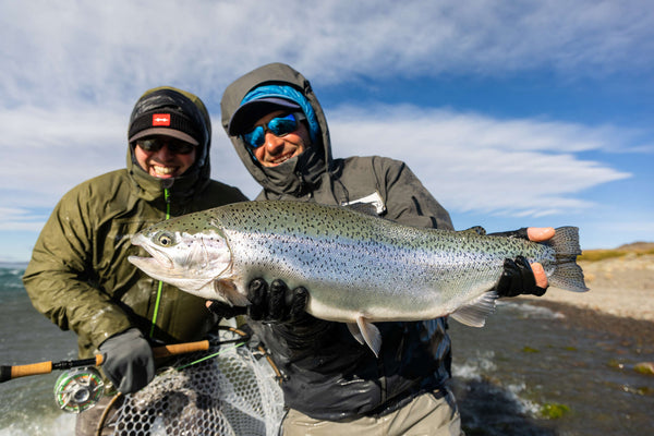 Two anglers in green and gray rain jackets holding a large silver rainbow trout on a fly fishing trip