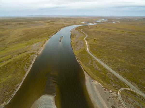 Aerial view of a winding river through flat green-brown tundra with a gravel road along the bank and overcast sky, trip