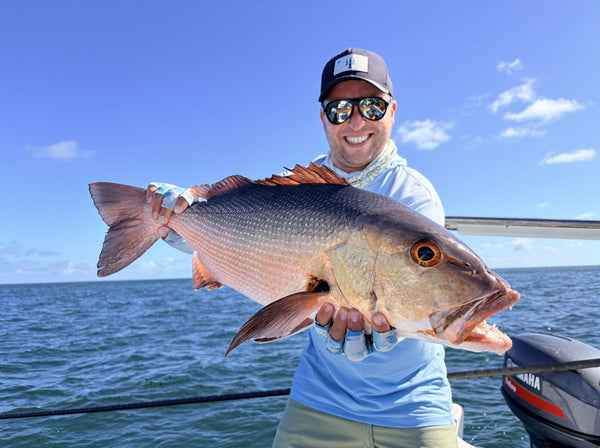 Smiling angler in navy cap and mirrored sunglasses holding large red snapper on boat under bright blue sky, fishing trip