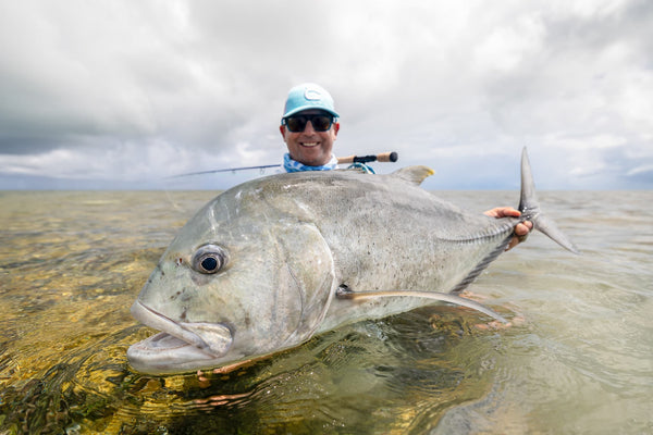 Angler in light blue cap holding large silvery trevally in shallow golden water under cloudy sky Providence Atoll fishing
