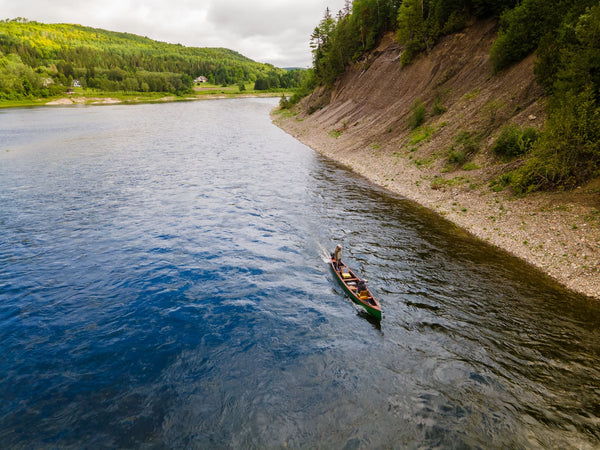 Two people paddling a green wooden canoe on deep blue Restigouche River beside a steep gravel cliff and lush forest
