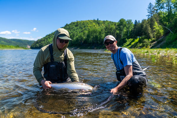Two anglers in olive hood and blue shirt wearing waders holding a large silver salmon in clear river with green forest banks