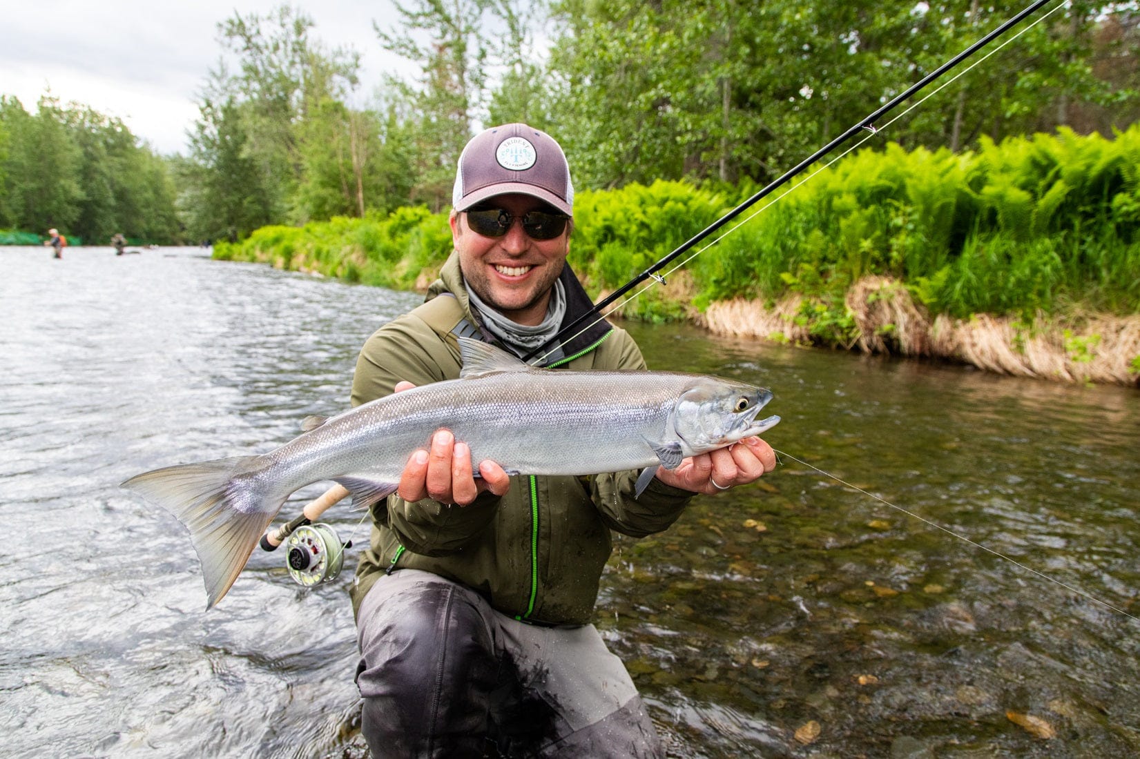 Smiling angler in olive jacket holding a silver trout over a shallow river with fly rod and reel, fly fishing gear visible