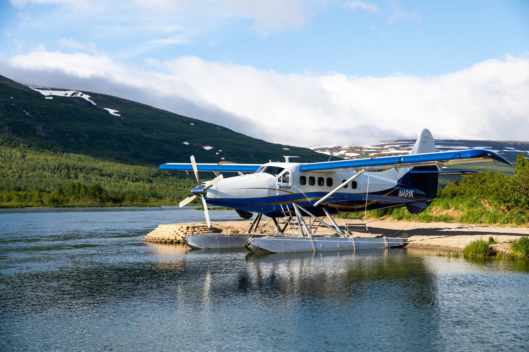 Blue-and-white floatplane with floats on calm Alaskan lake, green hills and snow patches — backdrop for Alaska trout flies